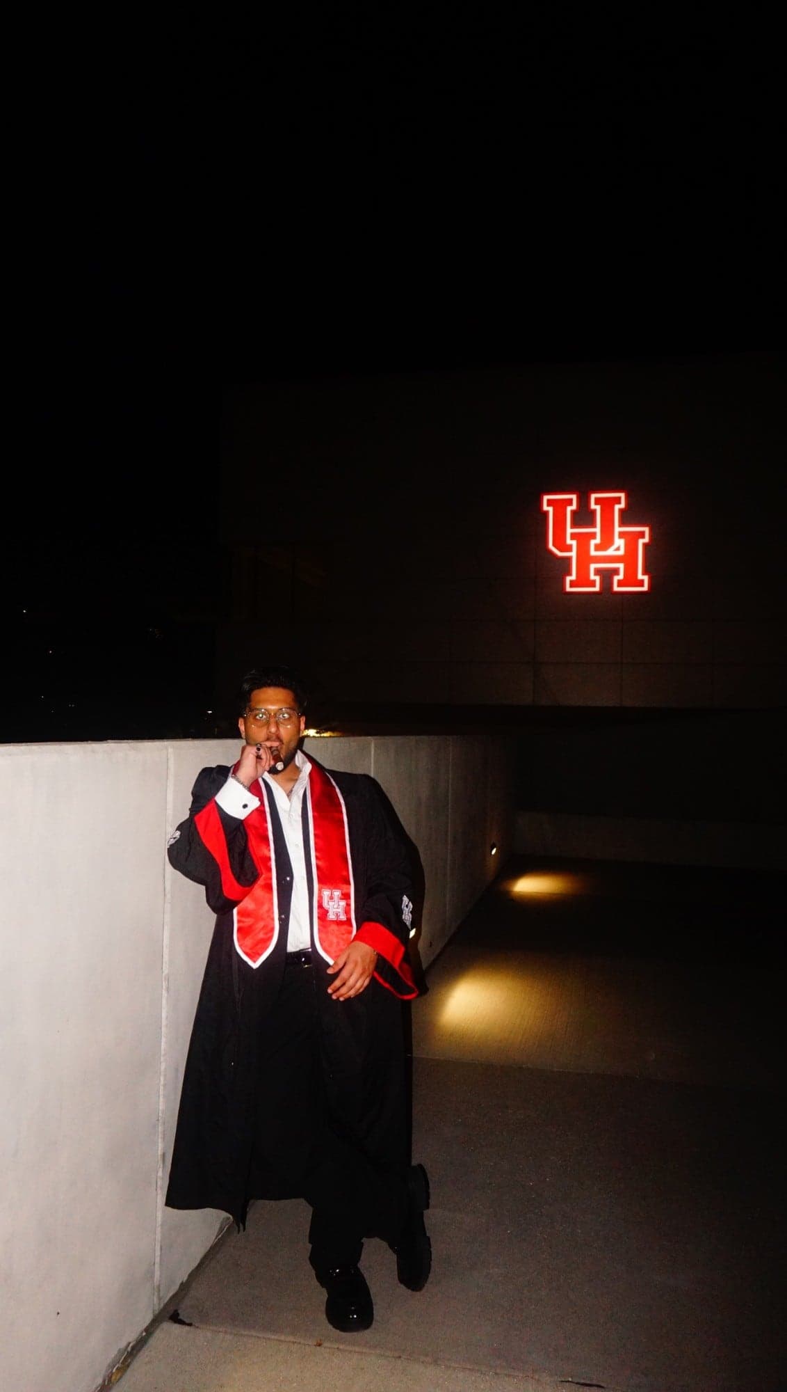 Luqman Ismat at night in a black graduation gown and red University of Houston stole, the UH logo glowing on the building behind him